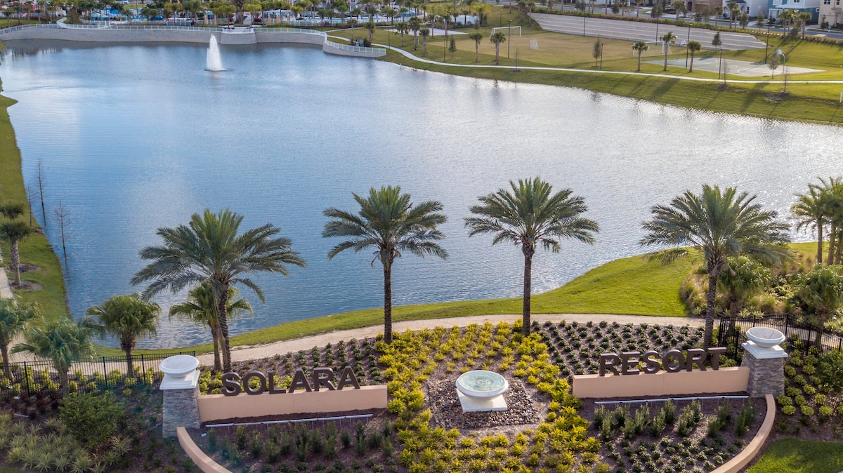 The entrance to Solara Resort is framed by palm trees and vibrant shrubs, creating an inviting landscape. A serene lake reflects sunlight, with a fountain gently cascading in the background. The resort's sign is prominently displayed, welcoming guests to the community.