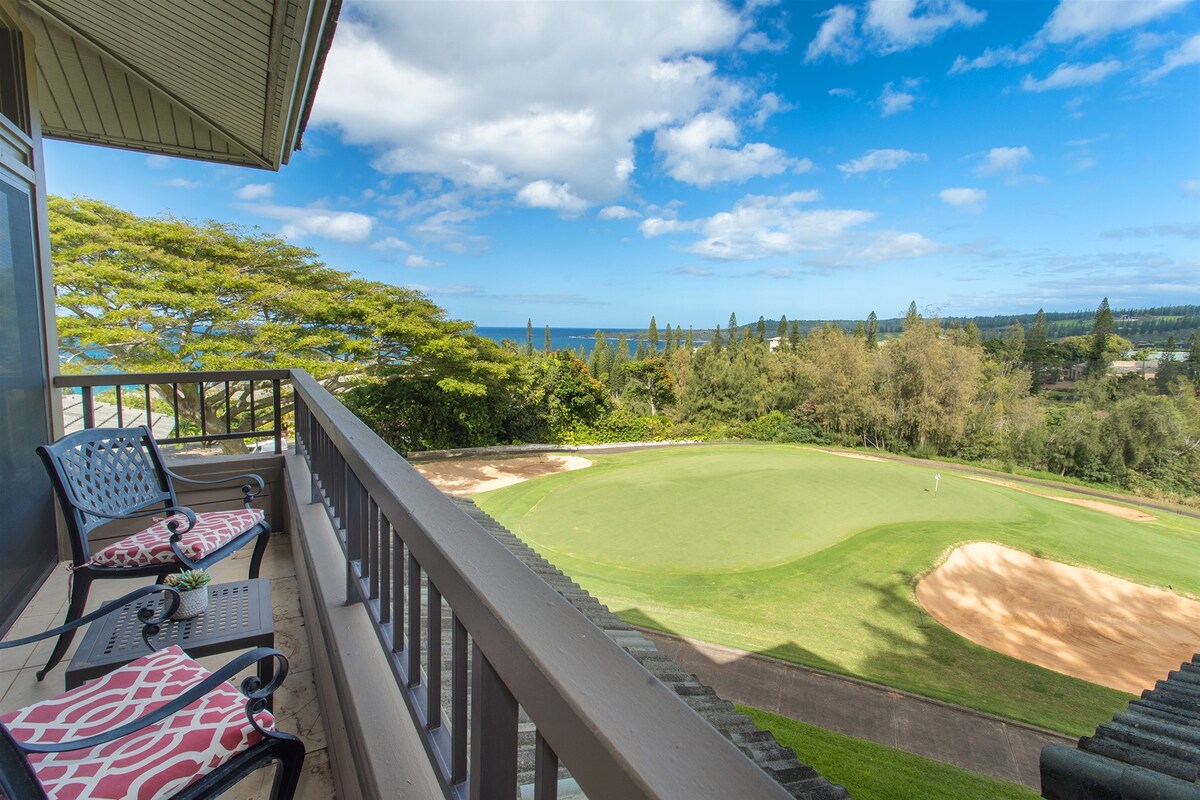 A spacious lanai overlooking the lush green golf course and the distant ocean. Two chairs with patterned cushions provide a comfortable spot to enjoy the view, framed by nearby trees and a clear blue sky.