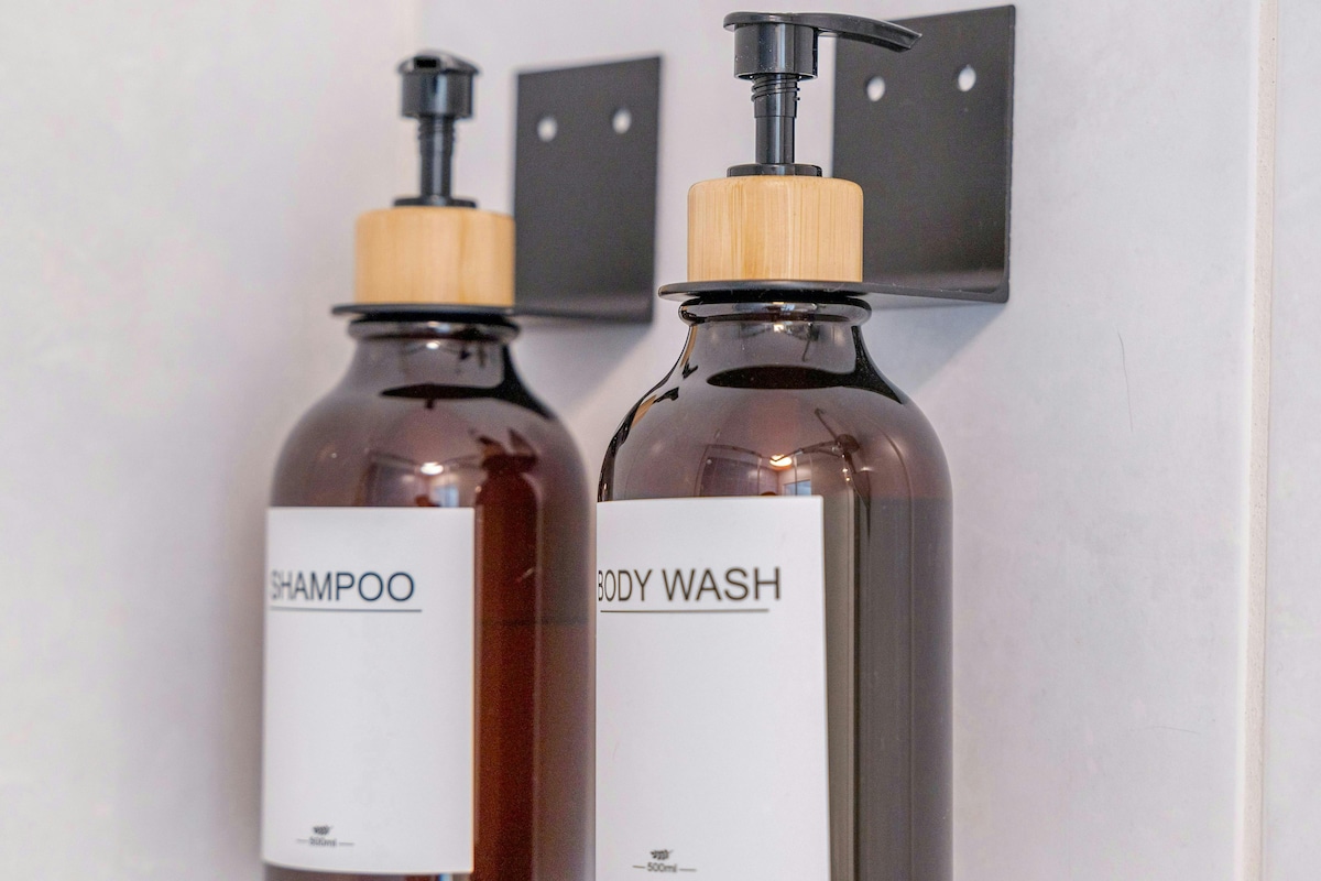 Two stylish wall-mounted dispensers are securely attached to a tiled bathroom wall. One dispenser is labeled 'Shampoo,' while the other is labeled 'Body Wash.' Both dispensers feature sleek dark brown bottles with wooden tops for easy access.