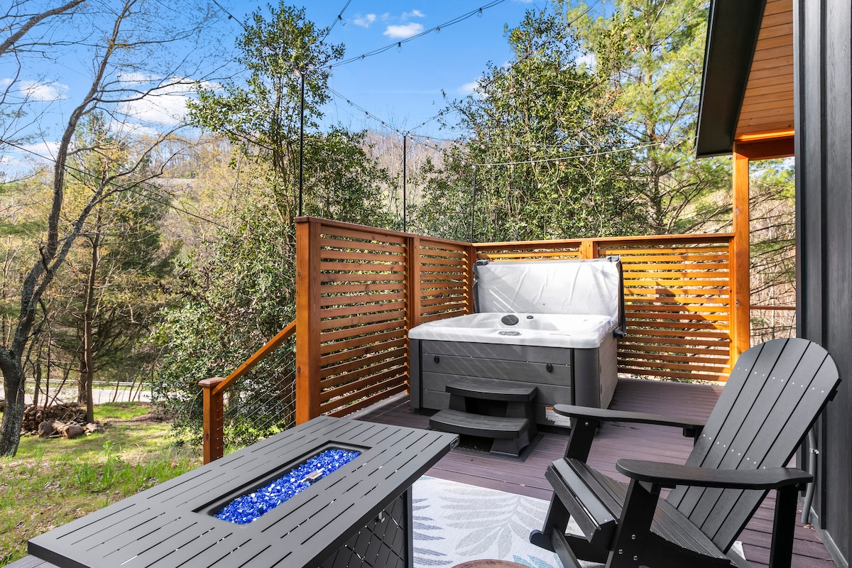 A private deck area features a hot tub surrounded by wooden slatted privacy panels and greenery. Two comfortable chairs are positioned beside a table with a blue stone surface. Soft lighting is provided by string lights overhead, creating an inviting outdoor space.