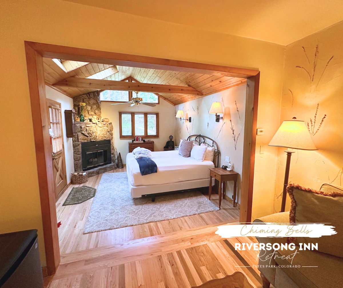A spacious suite is highlighted by a king bed positioned against a wooden accent wall. Natural light fills the room through large windows, while a stone fireplace adds warmth. The area features wooden flooring and soft textured rugs, creating a cozy retreat.