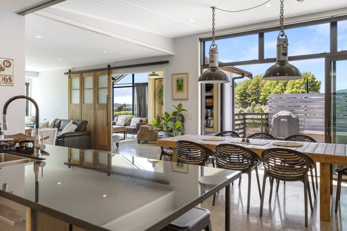 A contemporary kitchen area features a spacious island with a glossy countertop, surrounded by natural light from large windows. A dining table with stylish chairs is positioned adjacent to a cozy living space, enhancing the open-plan design. Decorative accents include plants and wall art, adding to the inviting atmosphere.