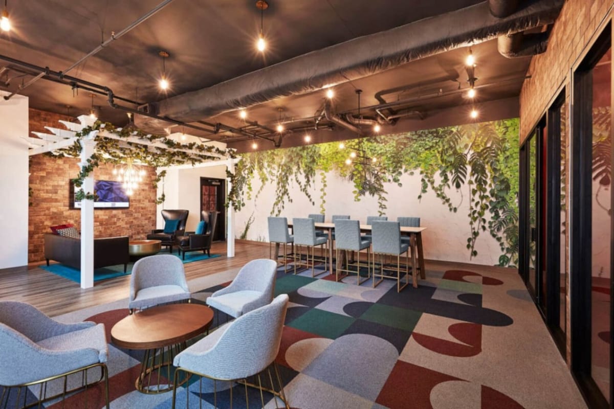 A modern shared space features a patterned carpet with various shapes and colors. Soft seating options are arranged around low wooden tables. A large mural depicting greenery adds a natural touch to the room, while warm lighting creates an inviting atmosphere.