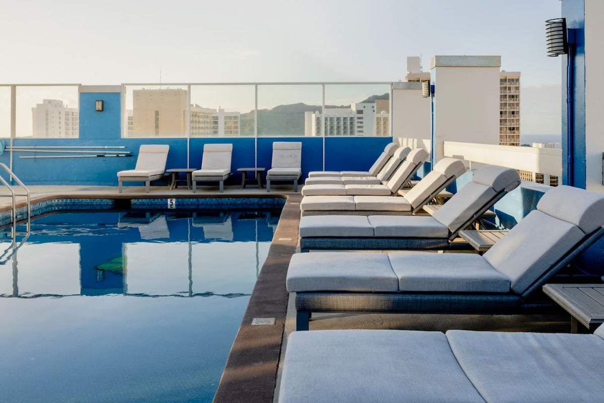A rooftop pool area features several lounge chairs arranged around a shimmering pool. The space is enclosed with glass railing, providing panoramic views of the surrounding cityscape. The water reflects the sky, and ample sunlight brightens the area.