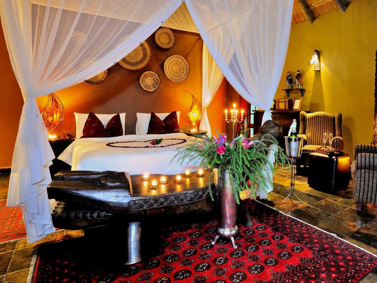 A serene sleeping area is framed by a delicate canopy, with a spacious bed draped in crisp linens. Decorative woven baskets adorn the wall, and a rich patterned rug covers the stone floor. Soft candlelight and fresh flowers enhance the tranquil ambiance.