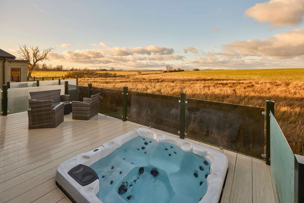 A private deck features a sunken hot tub surrounded by low glass panels. Comfortable outdoor seating is provided with two woven armchairs. The landscape reveals expansive fields under a serene sky, creating a peaceful outdoor setting.