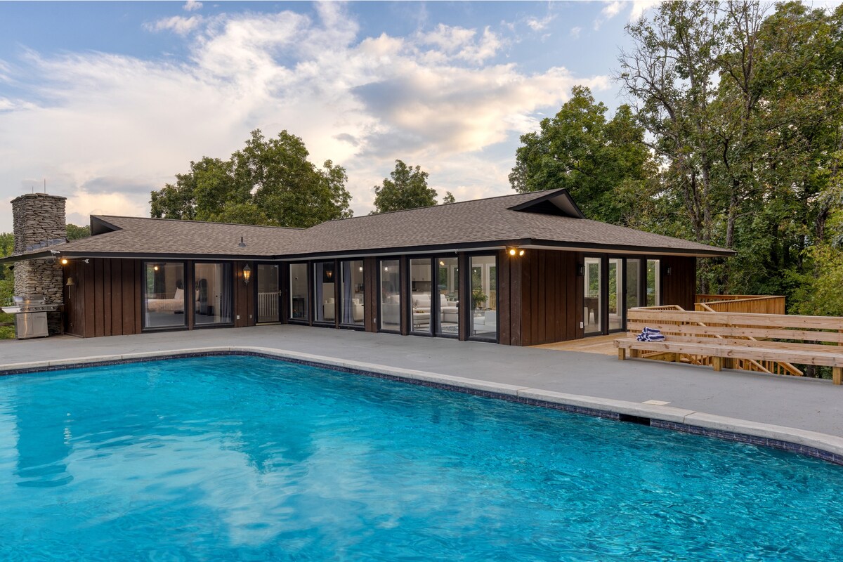The exterior of a modern retreat is displayed, featuring a spacious pool surrounded by a deck and large glass sliding doors. The building is set against a backdrop of lush trees under a partly cloudy sky, inviting moments of relaxation and leisure.