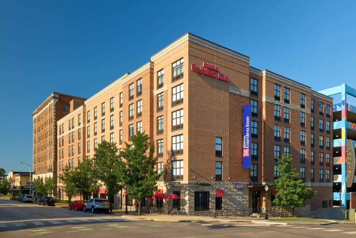 A modern hotel building with a brick facade showcases multiple levels and large windows. The entrance is marked by a small outdoor seating area with tables and umbrellas. Nearby, a colorful parking structure adds to the urban setting.