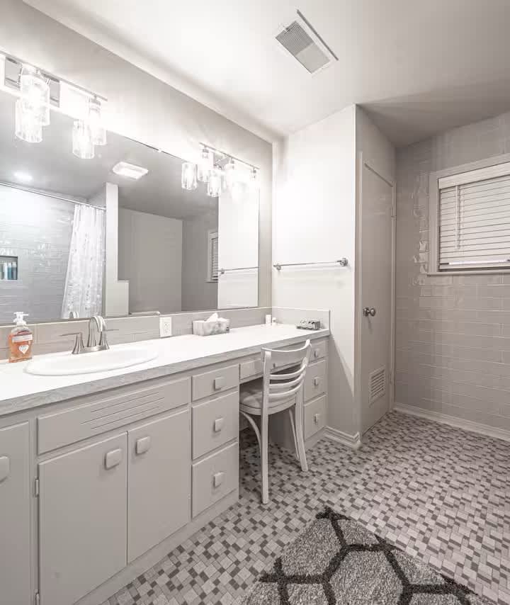 A bright bathroom features a large mirror above a spacious vanity with light fixtures. A white chair is positioned at a small desk area, while tile flooring in a geometric pattern adds texture. The room is well-lit, with a window featuring horizontal blinds.
