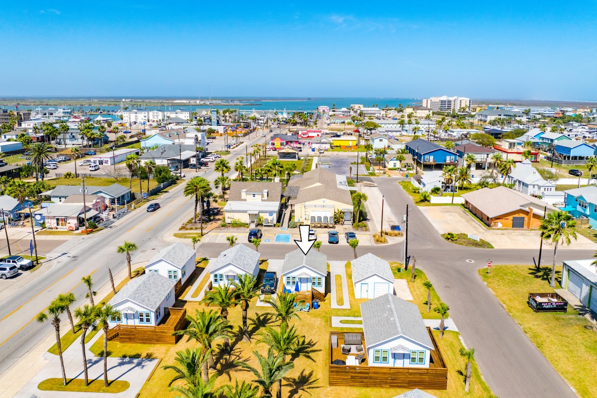 An aerial view showcases the property among a community of homes with vibrant roofs. Lush palm trees line the street, providing a tropical feel. A glimpse of the water is visible in the background, while the surrounding area features paved roads and a variety of buildings.