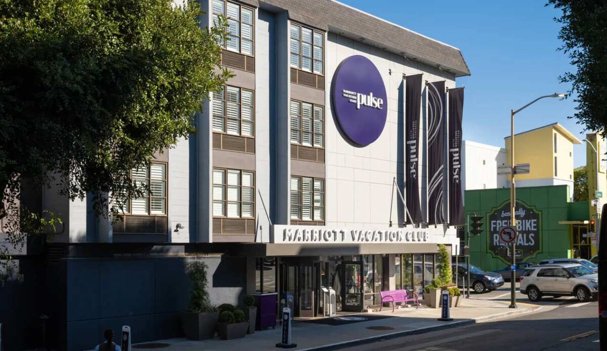The exterior of Marriott Vacation Club features a modern building design with large windows and a distinctive logo. The entrance is framed by a welcoming overhang, with a small outdoor seating area adorned in purple. Surrounding greenery adds a refreshing touch to the urban setting.