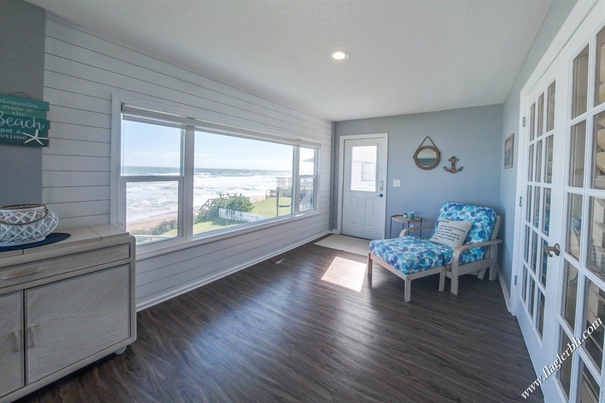 A bright sitting area features large bay windows showcasing ocean views and natural light. A comfortable blue-patterned lounge chair is present, along with a chest and wall decor reflecting a coastal theme. The room is painted in soft neutral tones, enhancing the serene ambiance.