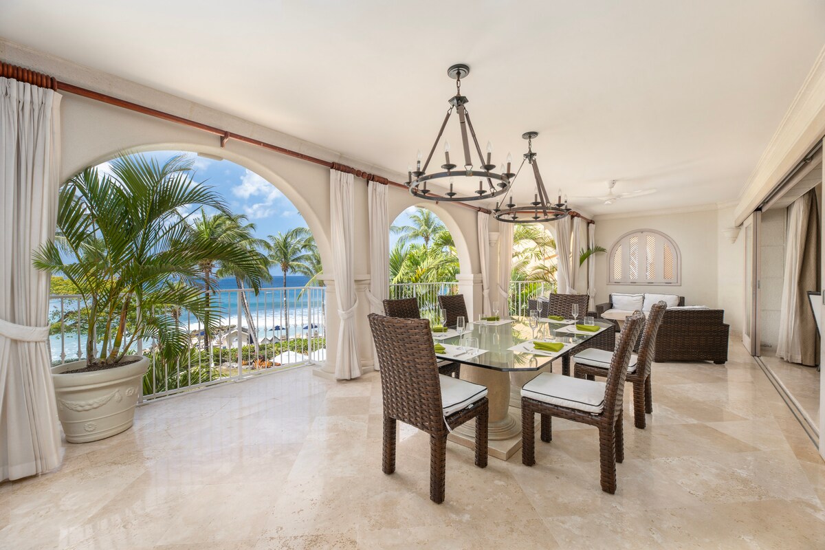 An inviting outdoor dining area features a large glass table surrounded by six woven chairs. The space is enhanced by tall palm trees and views of the turquoise Caribbean Sea through arched openings, complemented by soft light filtering through light curtains.