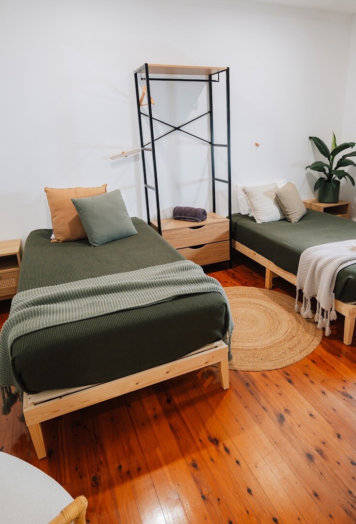 A bright and minimalistic guest room features two spacious beds adorned with soft green bedding. A wooden side table stands beside each bed, while a plant adds a touch of greenery. A modern shelving unit displays carefully arranged items against a neutral wall.