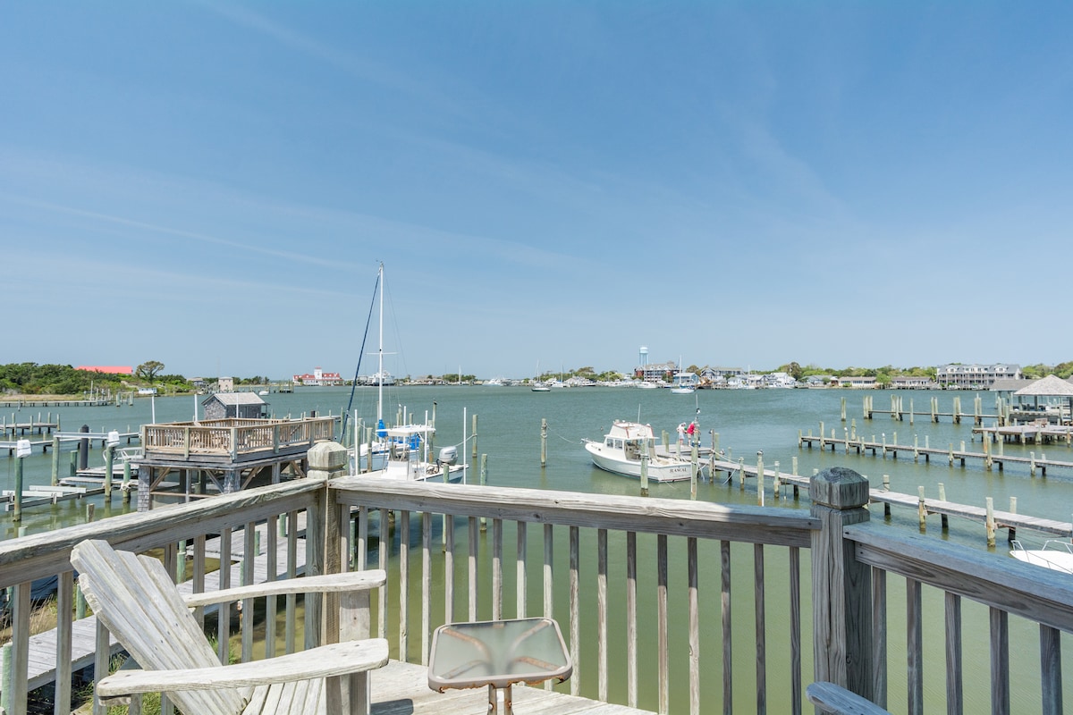 A serene waterfront view is captured from a wooden deck, featuring boats gently floating in the harbor. The scene includes a nearby sailboat and a scenic shoreline, framed by the surrounding docks and clear blue sky.