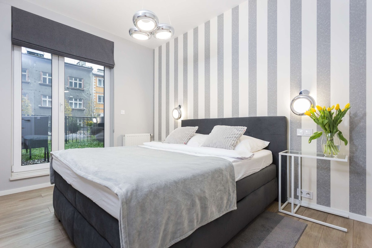 חדר שינה עם מיטה גדולה עם ראש מיטה אפור, שולחן קטן ליד המיטה עם אגרטל פרחים וקיר מבטא מפוספס. לוח הצבעים הניטרלי יוצר אווירה רגועה.