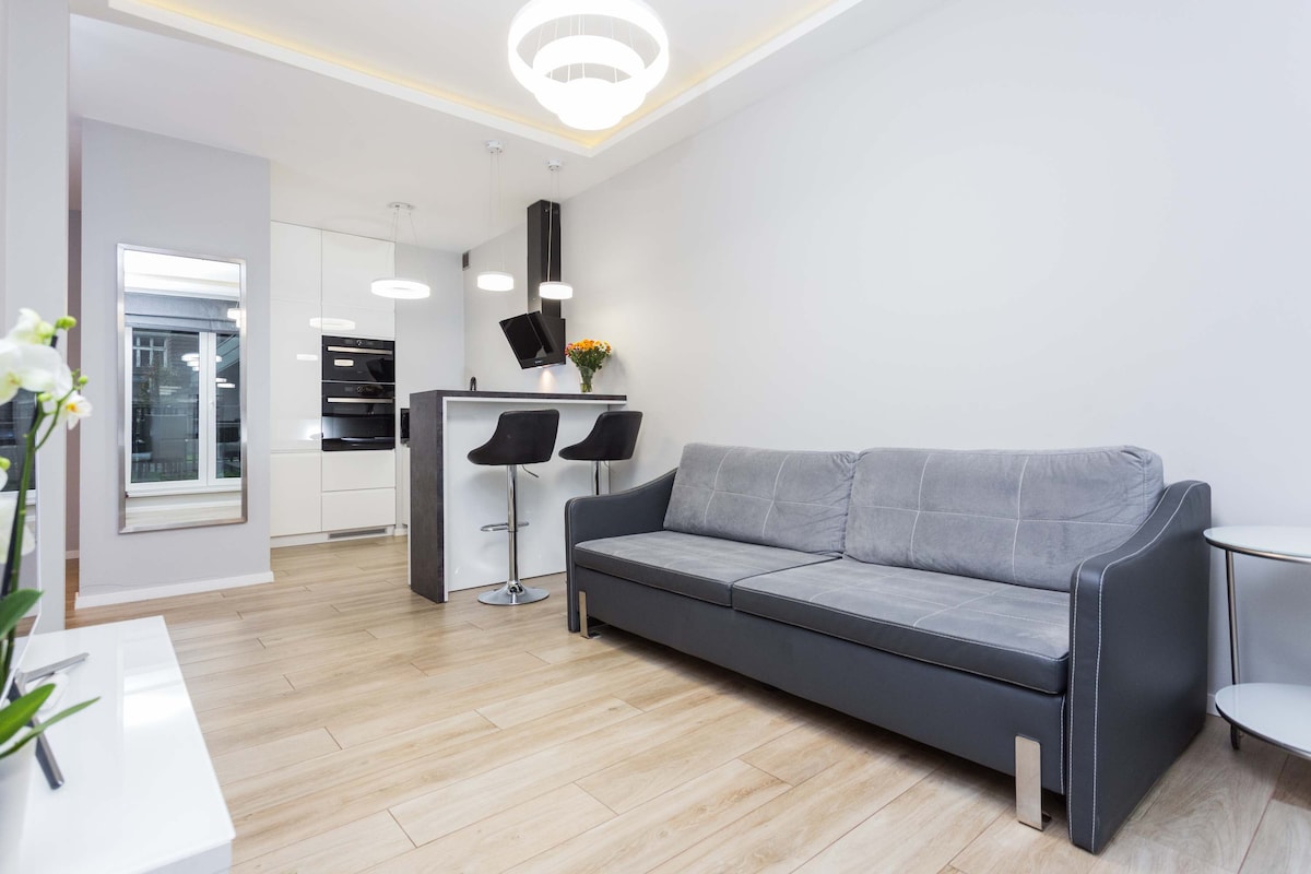 סלון מודרני עם ספה אפורה, דוכן טלוויזיה לבן ודלת זכוכית המובילה למרפסת. הנכס מואר באור טבעי.