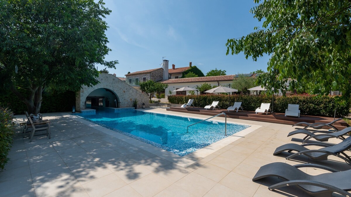 A heated outdoor swimming pool is centered in a spacious courtyard, surrounded by sunbeds for relaxation. Lush greenery and garden areas are visible in the background, along with an outdoor kitchen structure. A serene atmosphere is created by the clear blue sky and gentle sunlight.