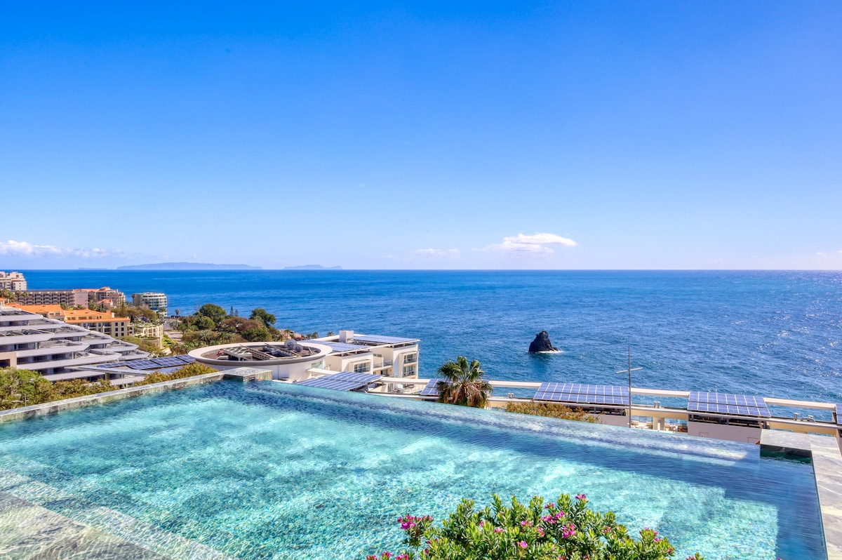 An infinity pool stretches towards the horizon, seamlessly blending into the expansive blue sea. A rocky outcrop is visible in the water, with lush greenery surrounding the pool area. The clear sky and gentle waves enhance the tranquil setting.