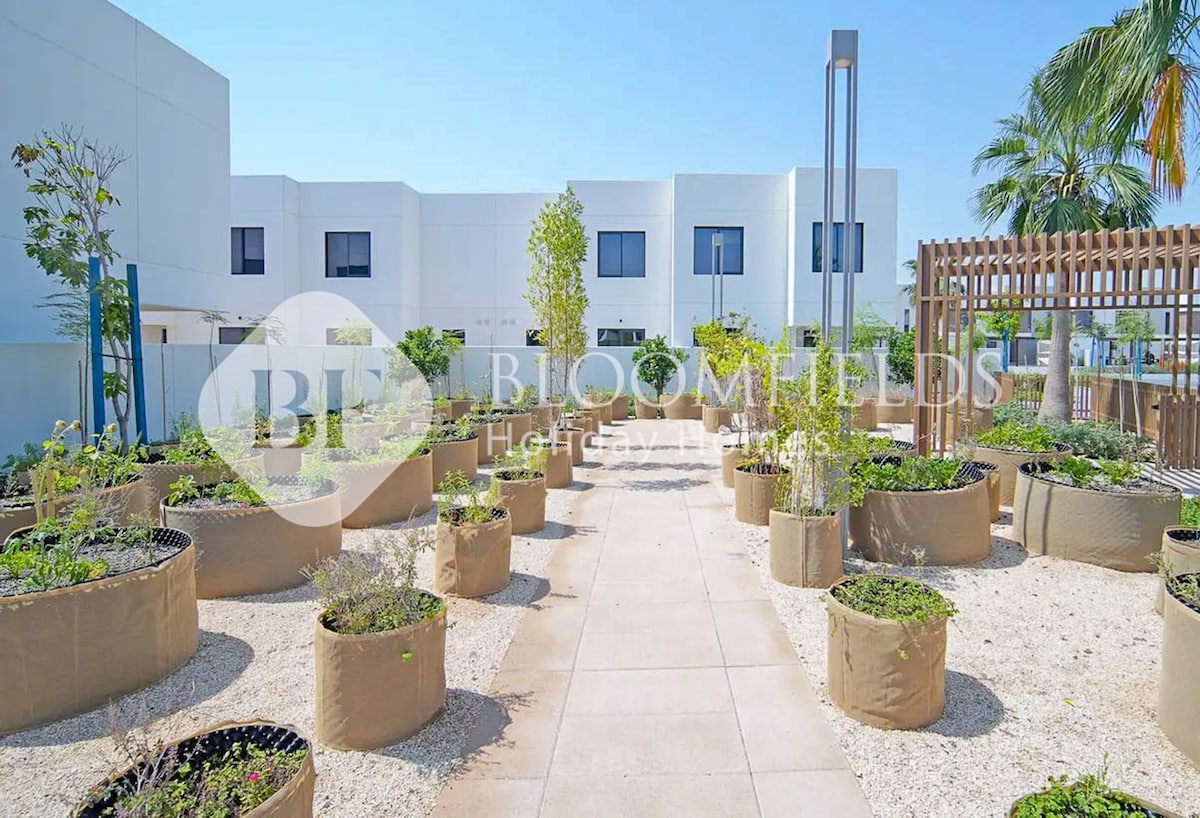A sunlit outdoor garden area features several round planters filled with lush greenery. The pathway, made of light-colored stone, leads through the garden, surrounded by low walls. Modern buildings are visible in the background, with palm trees adding to the serene atmosphere.