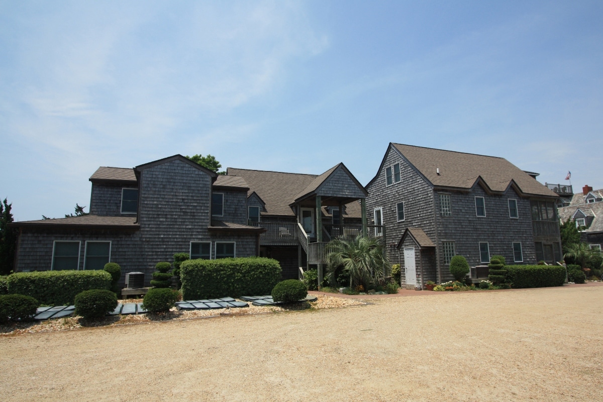 A two-story villa is seen, featuring a combination of dark shingles and multiple gabled roofs. Surrounding landscaped areas include manicured shrubs and pathways leading to the entrance. A spacious parking area is visible in front, enhancing access to the building.