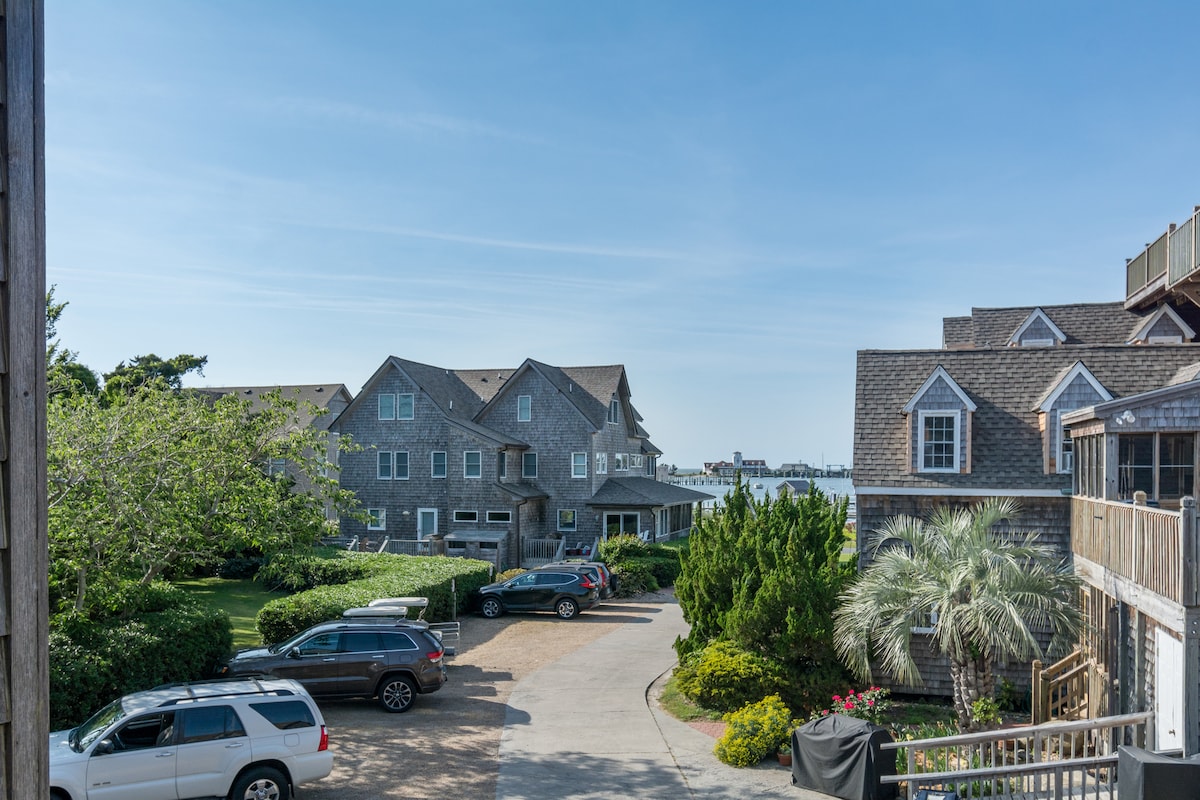 The image showcases a view from the balcony, highlighting a landscaped area with multiple trees and shrubs. A pathway leads towards the parking area, where several vehicles are visible. In the distance, the outline of structures can be seen, suggesting a coastal setting.