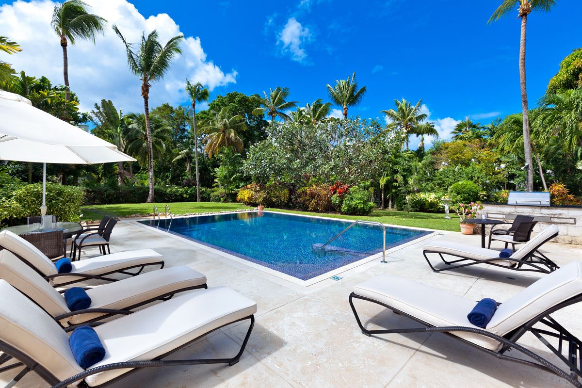 The outdoor pool area is surrounded by lush tropical gardens, featuring a tranquil saltwater pool with loungers positioned nearby. An umbrella provides shade, while palm trees and vibrant foliage enhance the serene setting under a clear blue sky.