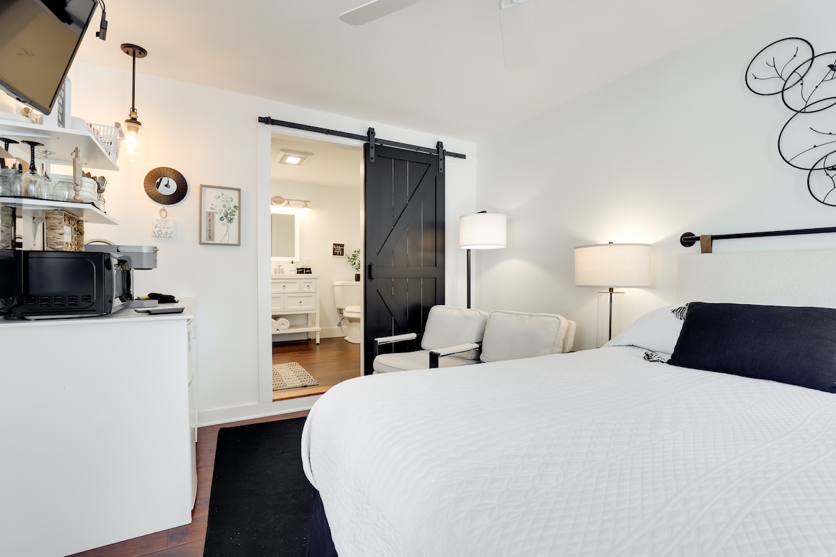The studio features a spacious bed with a textured white coverlet and a decorative wall art piece above. A cozy seating area with two chairs is situated near a kitchenette, which contains a microwave and other essentials. A sliding barn door leads to the bathroom, enhancing the modern design.