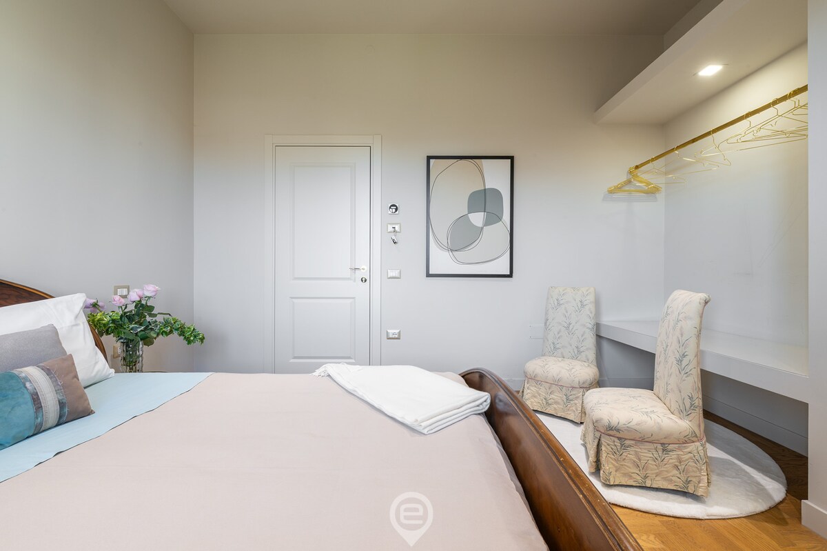 A spacious room features a double bed with neutral bedding and decorative pillows. Two light chairs are positioned beside a small table. A wardrobe with hanging space is visible, along with a minimalistic art piece on the wall. Natural light illuminates the area.