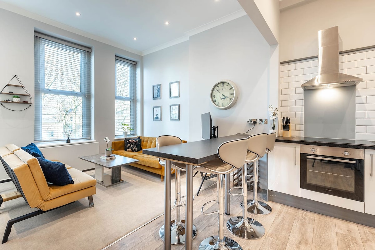 A light-filled living area features a mix of comfortable seating, including a stylish yellow sofa and a sleek gray sofa bed. A modern kitchen is visible, equipped with a stove, oven, and ample countertop space. Two large windows allow natural light to enhance the atmosphere.
