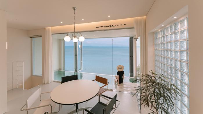A dining area features a round table surrounded by minimalist chairs. Large glass walls provide unobstructed views of the ocean, while soft curtains frame the windows. Natural light fills the space, enhancing the calm atmosphere created by the soft color palette.