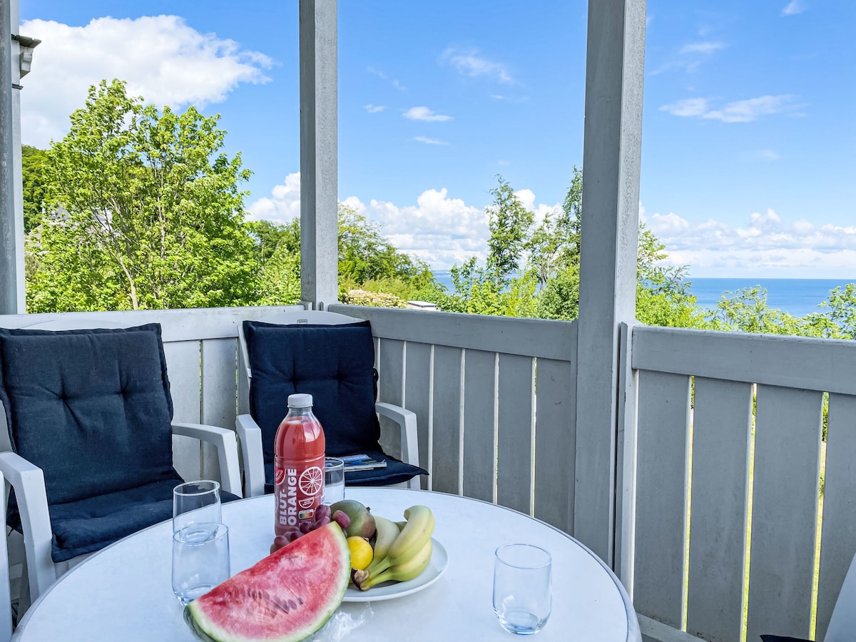 A balcony setup is featured with a round table and two chairs, surrounded by lush greenery. Fresh fruit, including watermelon and bananas, is arranged on the table next to a bottle of beverage and glasses. A scenic view of the sea is visible in the background under a clear sky.
