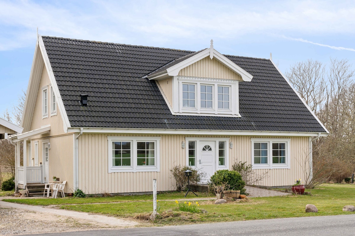 A spacious two-story holiday home with a light-colored wooden facade and a black tiled roof is portrayed. Large windows enhance natural light, while a welcoming front door is framed by flower beds. A gravel path leads to the entrance, surrounded by a manicured landscape.