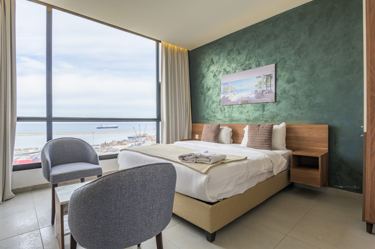 A spacious room showcases large windows offering panoramic sea views. A double bed with crisp white linens is complemented by a wooden bedside table. Two armchairs and a small table create a cozy seating area, framed by soft drapes that gently filter natural light.