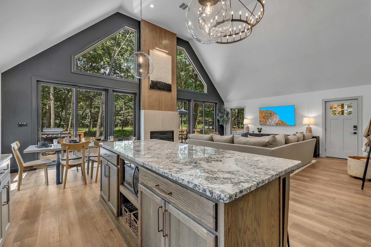 An open-concept living area showcases a modern kitchen island with a gray granite countertop. Bright windows allow natural light to fill the space, where comfortable seating is arranged near a gas fireplace. A dining table is visible, alongside outdoor views through expansive glass walls.