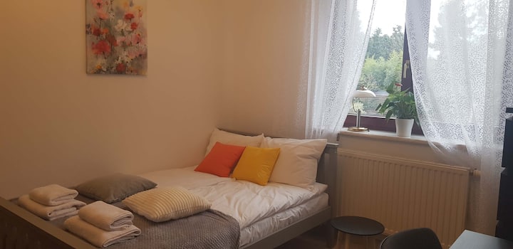 غرفة نوم بسرير مغطى ببطانية ناعمة ووسائد برتقالية. تسمح النافذة ذات الستائر البيضاء بدخول الضوء الطبيعي.