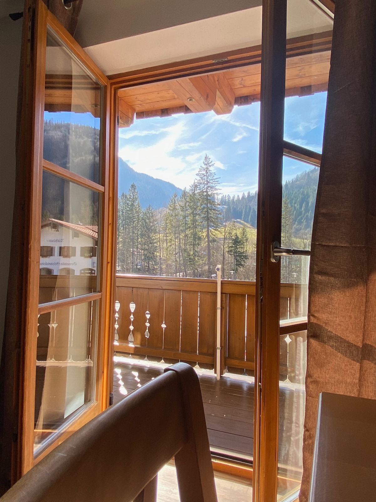 A French door opens to reveal a private balcony, framed by natural wood. Scenic views of lush green trees and distant mountains are visible under a bright sky. Sunlight creates a warm ambiance within the space, emphasizing the inviting connection to the outdoors.