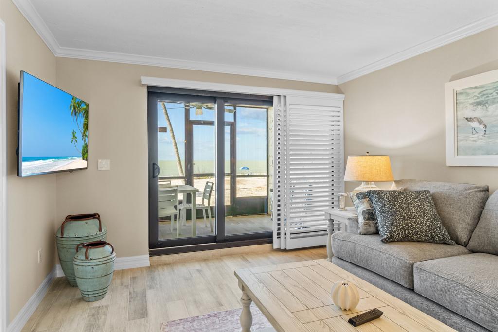 A light-filled living area is presented, featuring a comfortable gray sofa and a wooden coffee table. Sliding glass doors lead to a light-filled patio with views of the beach. Decorative elements include a framed art piece on the wall and a flat-screen TV mounted nearby.