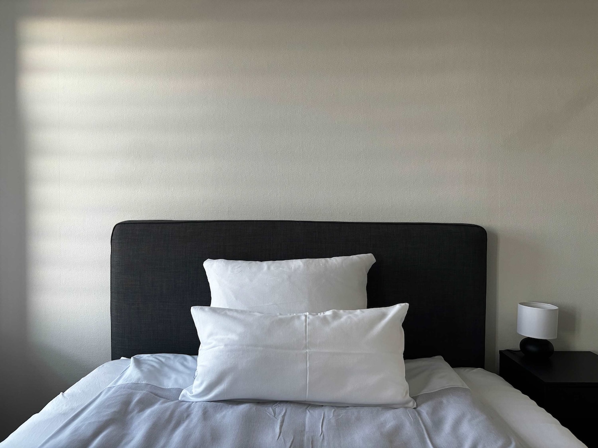 A spacious bed is featured with a dark gray headboard and two neatly arranged white pillows. Soft light filters through nearby window coverings, illuminating the bedding. A small bedside lamp sits on a dark nightstand, adding a simple touch to the cozy sleeping area.