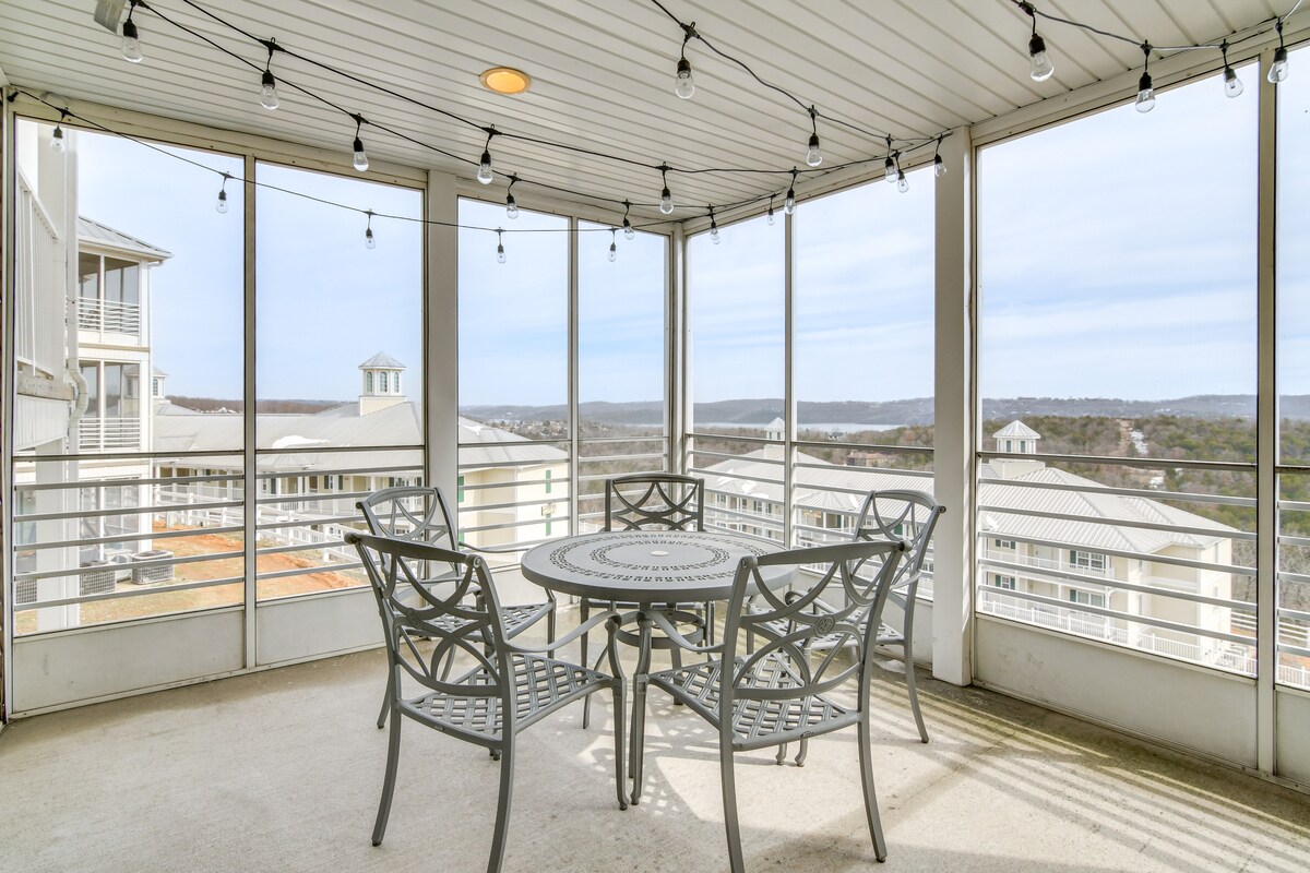 A screened-in balcony features a round dining table surrounded by six metal chairs. String lights are hung overhead, enhancing the space. Expansive views of the surrounding landscape and distant hills can be observed through the large glass panels.