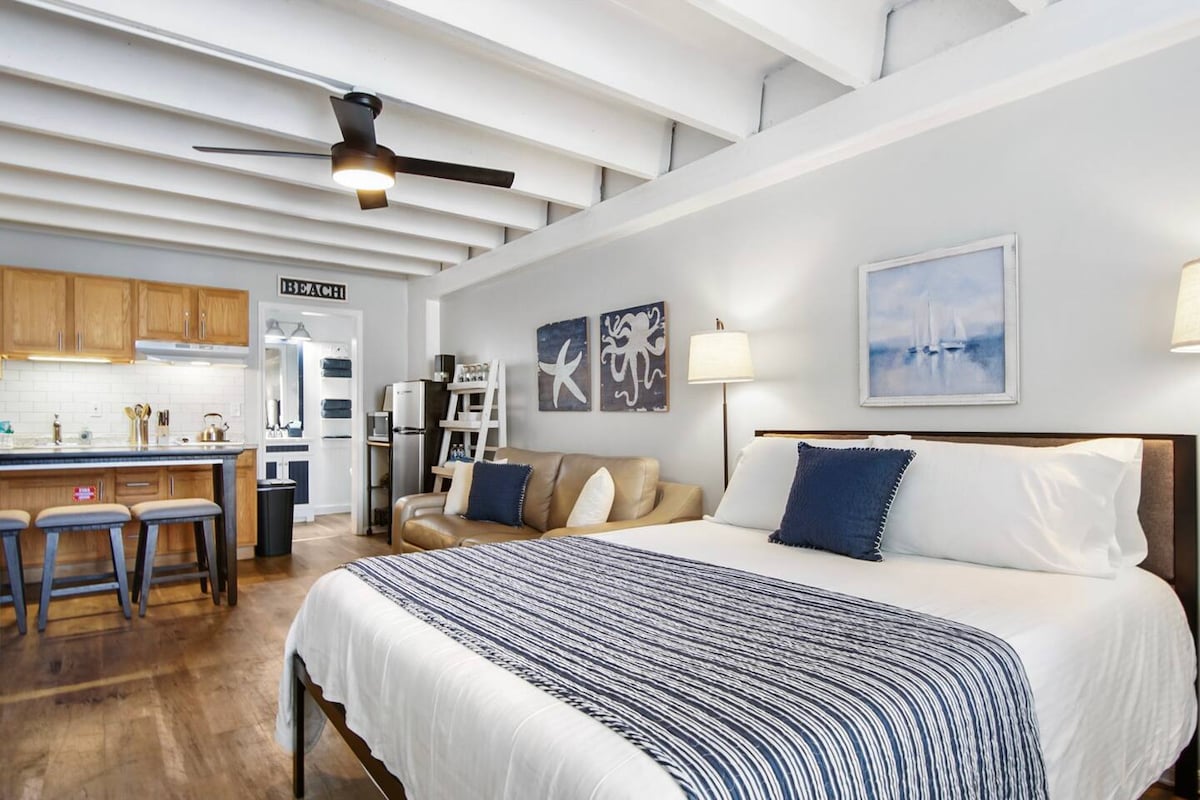 A spacious room features a large bed with white linens and a navy striped blanket. A comfortable sofa is positioned nearby. Soft lighting from two lamps complements the natural light streaming through the window. The kitchenette is visible in the background, showcasing wooden cabinets and modern appliances.
