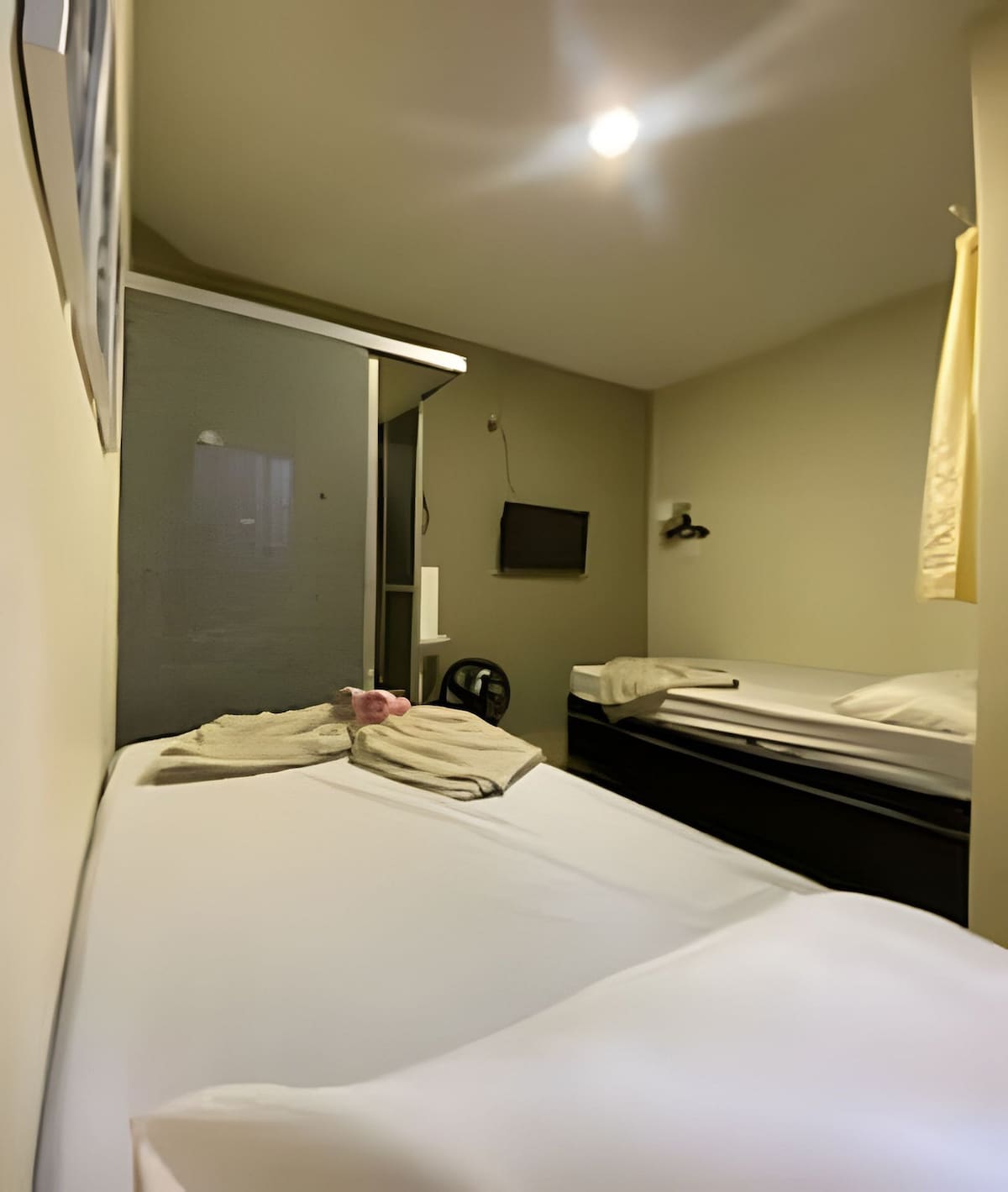 A cozy sleeping area is presented with two beds, one made up with fresh linens and towels placed neatly on top. A wall-mounted television is visible, and light illuminates the neutral-toned walls, creating a calm and comfortable environment.