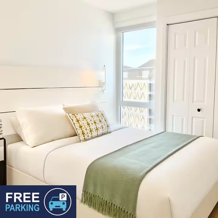 A well-lit sleeping area features a spacious bed adorned with crisp white linens and a decorative throw pillow. A green throw blanket lies across the foot of the bed. Natural light enters through a large window, enhancing the bright, clean ambiance of the room.