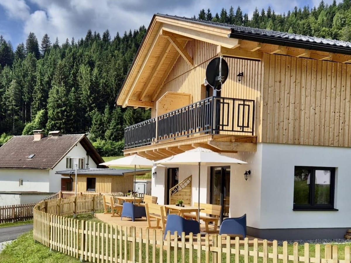 The semi-detached chalet features a wooden exterior with a small balcony overlooking the surrounding greenery. A spacious terrace is furnished with outdoor seating, and a wooden fence surrounds the property, enhancing its rural charm. Tall trees are visible in the background.