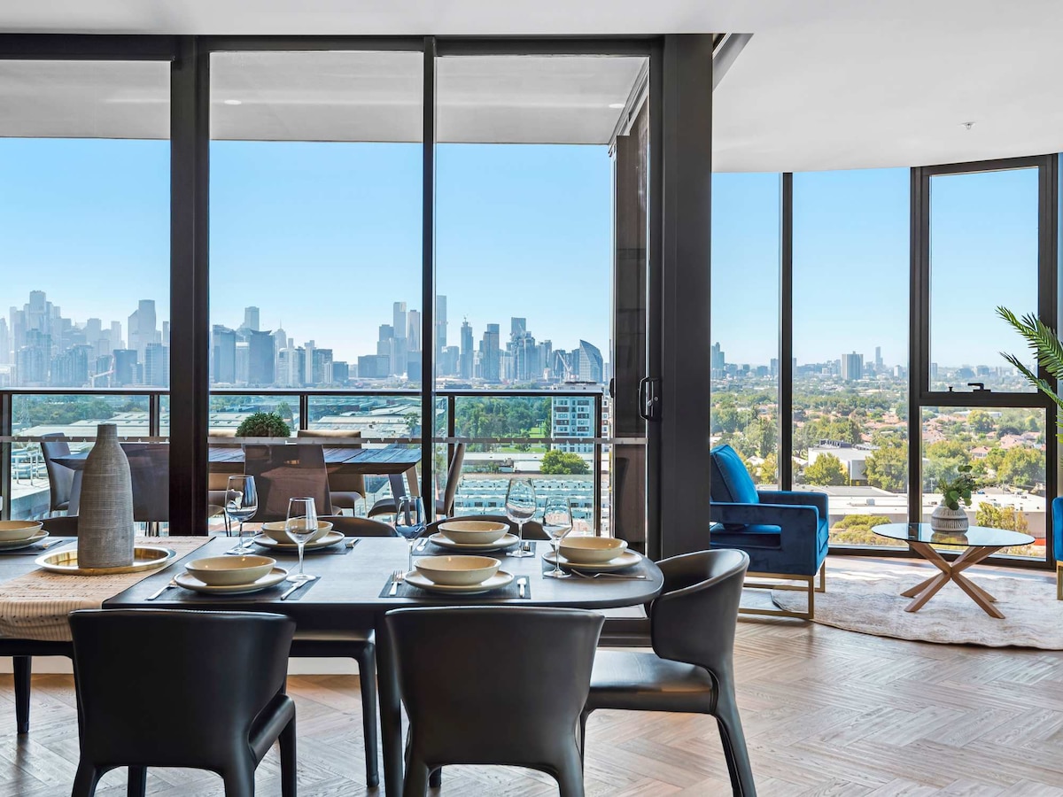 A dining area features a sleek table set for six, accompanied by stylish black chairs. Floor-to-ceiling windows showcase panoramic views of Melbourne's skyline, allowing natural light to fill the space. The room is elegantly furnished with modern decor and a small green plant adds a touch of nature.