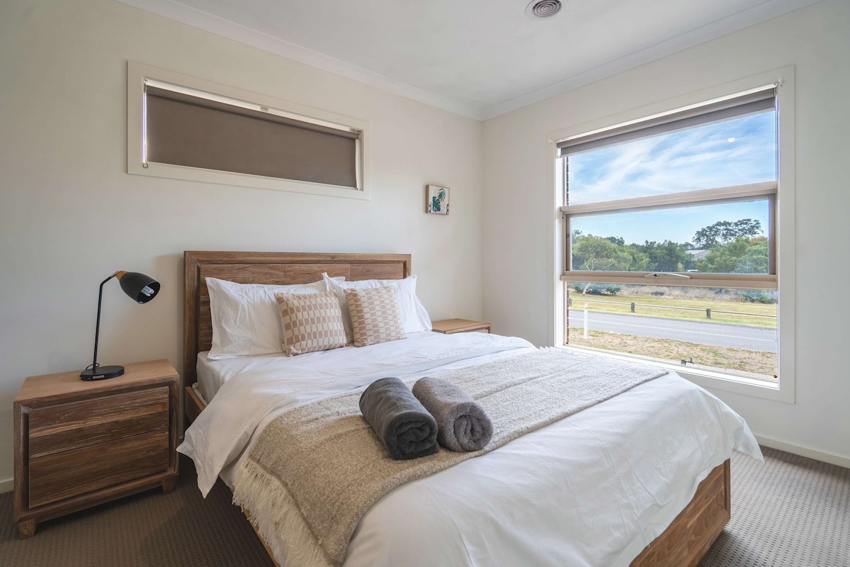 A well-appointed bedroom features a spacious bed with white bedding and decorative cushions. Two rolled towels are neatly placed at the foot of the bed. A bedside table holds a lamp, and large windows allow natural light, offering views of the outdoors.