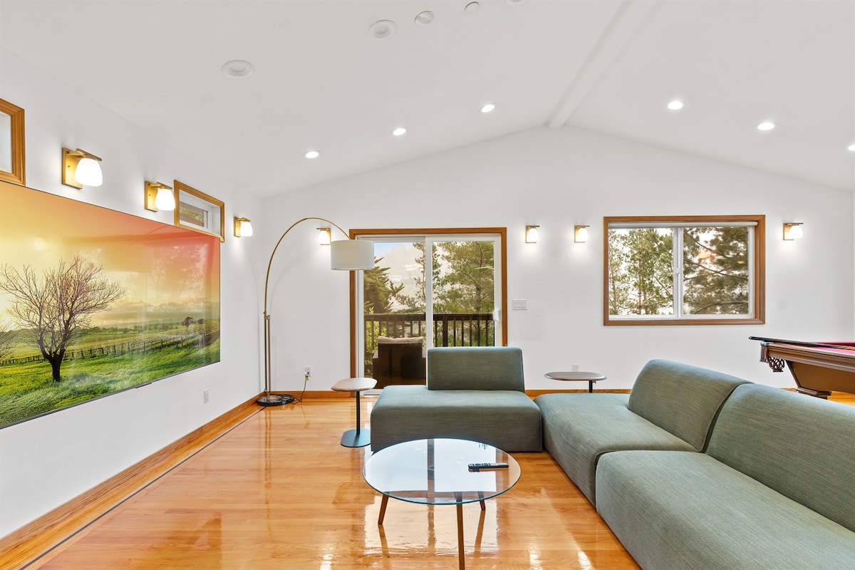 The open living area features a modern sectional sofa arranged around a glass coffee table. A floor lamp provides additional lighting, and large windows allow natural light to fill the space. A vibrant landscape artwork adorns one wall, enhancing the overall ambiance.