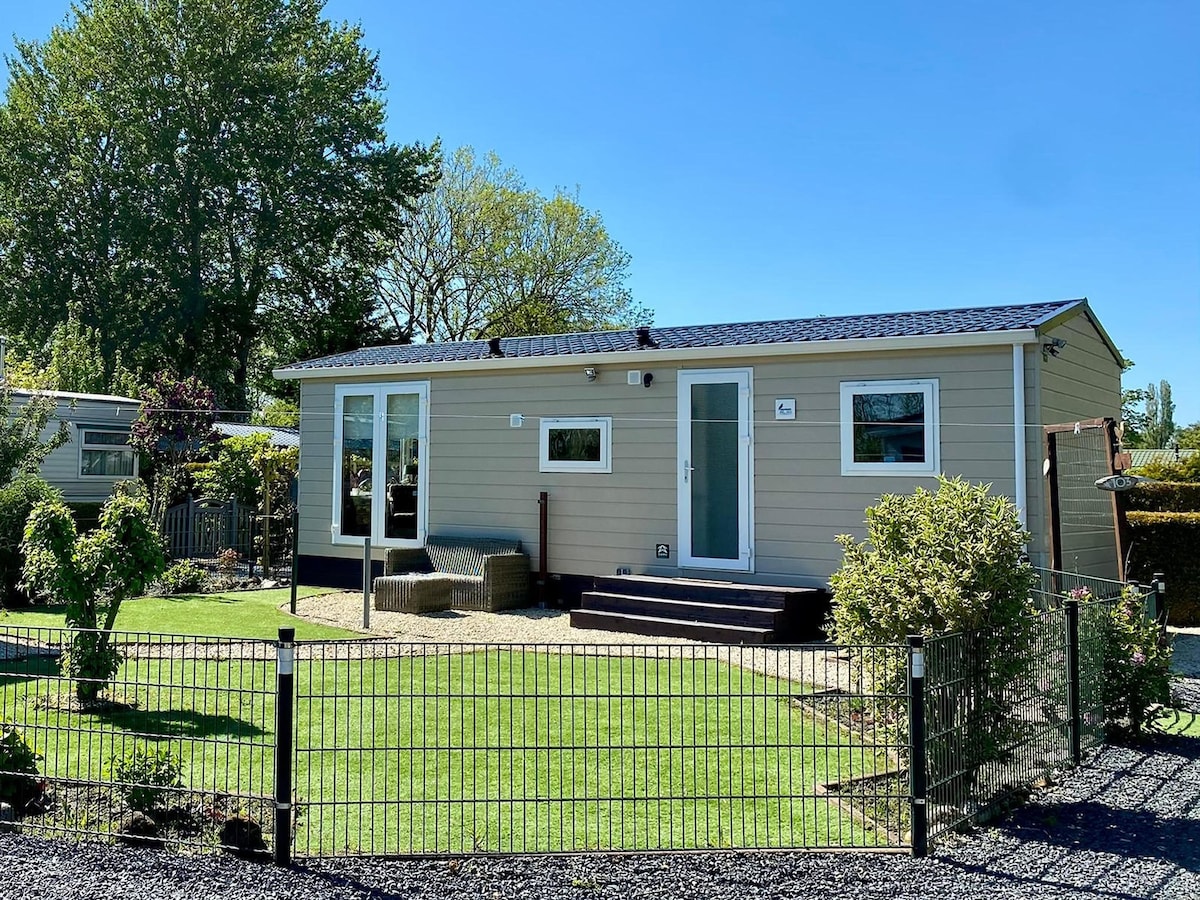 A modern chalet is surrounded by a well-maintained garden featuring a lush green lawn and ornamental shrubs. The structure includes large windows and a front porch with steps leading to the entrance. Fencing encloses the property, offering a sense of privacy.