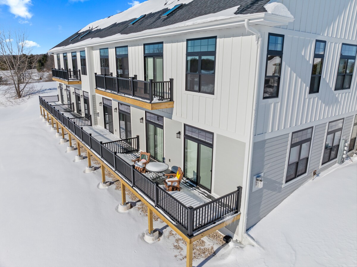 The exterior of the newly constructed building is shown, featuring a row of balconies with black railings and comfortable seating. Snow blankets the ground, creating a serene backdrop for the modern facade, characterized by large windows that allow ample sunlight into the residences.