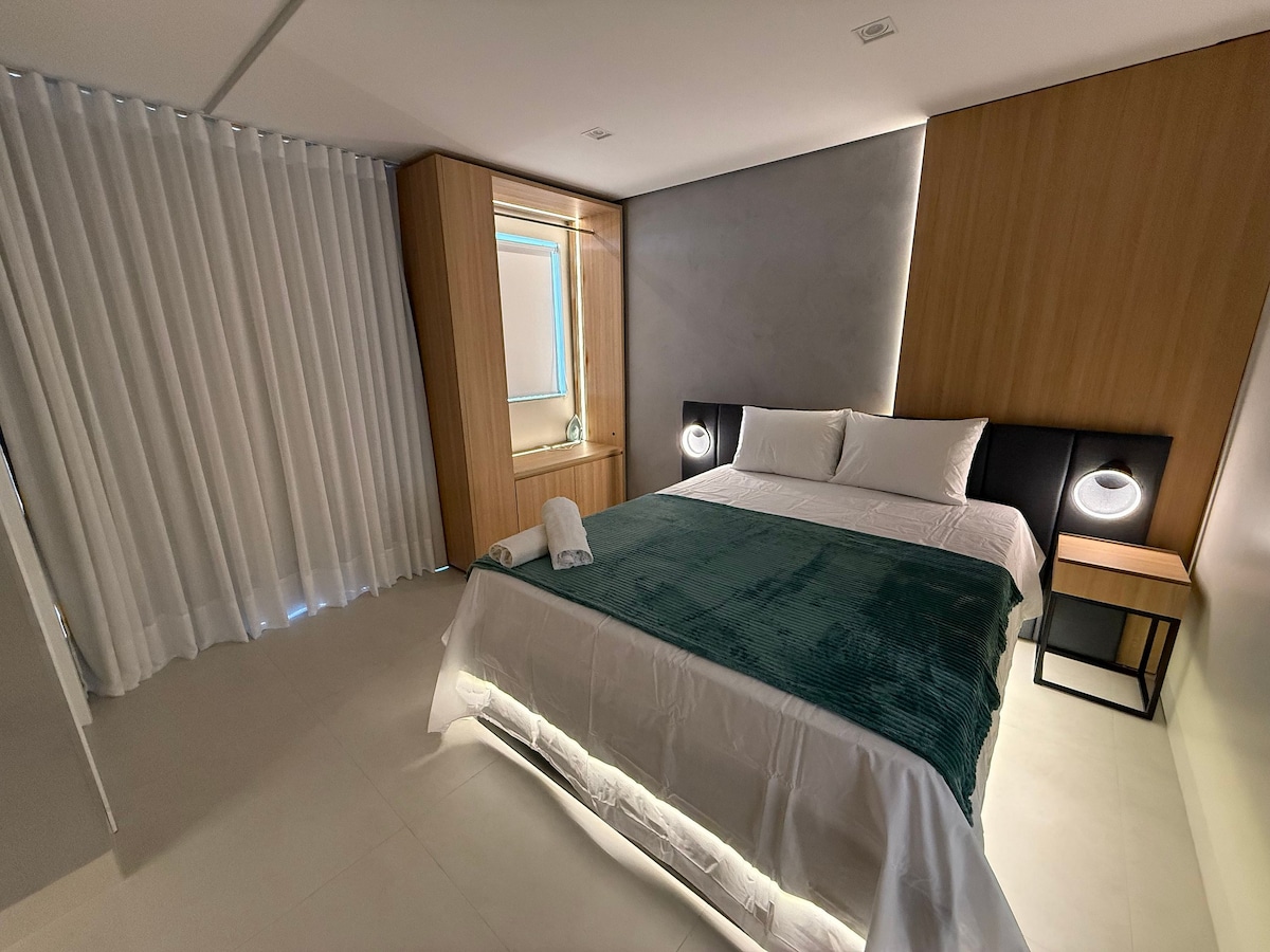 The bedroom features a spacious bed covered with a soft green throw. Minimalist wooden cabinetry and modern lighting create a calming aesthetic. Large, light-filtering curtains frame a window, while bedside tables are positioned on each side of the bed, adding convenience and style.